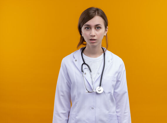 穿惊讶的年轻女医生穿着医用长袍和听诊器站在隔离的橙色墙上 有复印空间年轻人惊喜女人