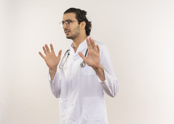 医生神经质的年轻男医生 戴着眼镜 穿着白色长袍 手持听诊器 在隔离的白色墙壁上举手 还有复印空间年轻吱吱穿