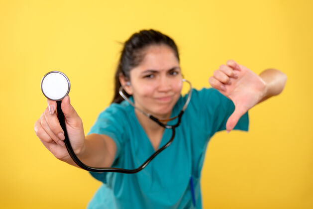 做黄色墙上年轻女医生手持听诊器做拇指朝下标志的正面图下医疗年轻女医生