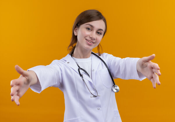 年轻面带微笑的年轻女医生穿着医用长袍 手持听诊器 在孤立的橙色墙上伸着手手女人医生