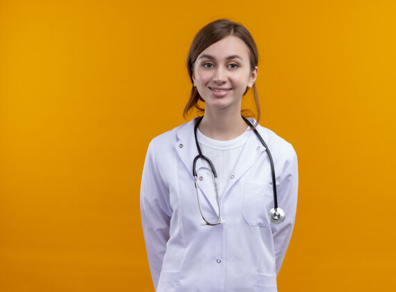 年轻面带微笑的年轻女医生穿着医用长袍和听诊器 双手背在身后 站在隔离的橙色墙壁上 有复印空间穿听诊器女人