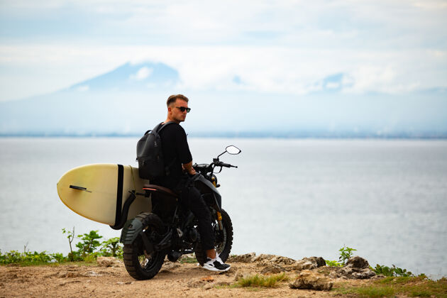 家伙年轻人坐在摩托车上 手里拿着冲浪板探索海滩冲浪板