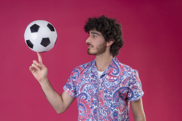 足球年轻英俊的卷发男人用手指拿着足球 看着孤立的粉红色墙上的足球男人抱着年轻