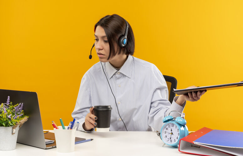 剪贴板专注的年轻呼叫中心女孩戴着耳机坐在办公桌旁拿着剪贴板和塑料咖啡杯 看着孤立在橙色上的笔记本电脑杯子耳机年轻
