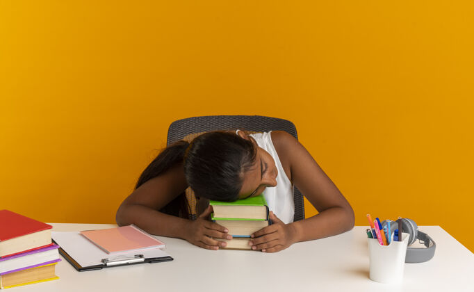 书桌年轻的女学生坐在书桌旁 手里拿着工具 把头放在橘色墙上孤立的书本上女学生年轻头