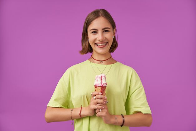 项链女学生 双手拿着冰激凌的年轻女士 露出舌头 对着镜头微笑 越过紫色的墙壁穿着绿色的t恤 手镯 戒指和项链牙套面部女士