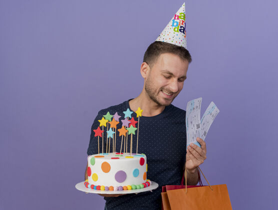 机票戴着生日帽的白种人帅哥拿着生日蛋糕纸购物袋礼盒和机票 背景是紫色 有复印空间包礼品机票