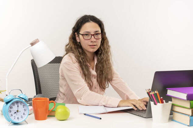 笔记本电脑自信的年轻漂亮的女学生戴着眼镜坐在书桌旁 拿着学习工具 用白色的笔记本电脑做作业工具坐着女学生