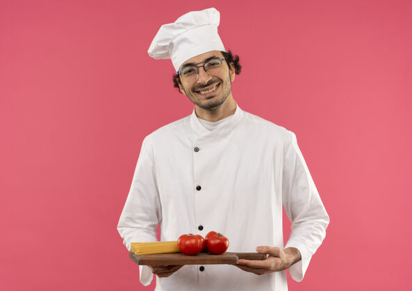男年轻的男厨师穿着厨师制服 戴着眼镜 手里拿着西红柿意大利面 放在粉红色的墙上请玻璃杯拿着