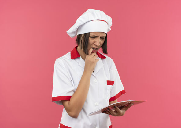 复制困惑的年轻女厨师穿着厨师制服 拿着手指头看着笔记本 嘴唇上孤立着粉色的复印空间制服女性厨师