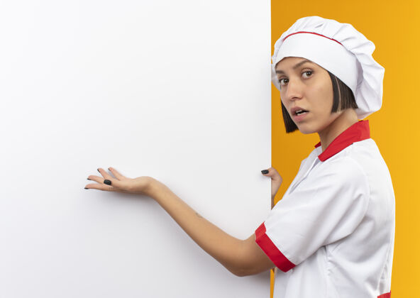 墙壁穿着厨师制服的年轻女厨师站在白色的墙壁旁 手拿着并用手指着隔离在橘黄色空间里的厨房空间指向烹饪