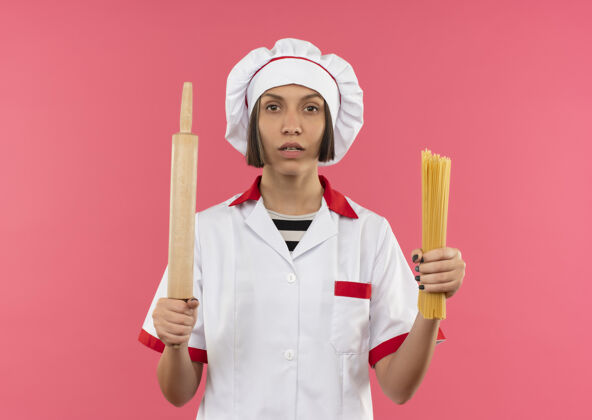 制服自信的年轻女厨师身着厨师制服 手里拿着意大利面和擀面杖 穿着粉红色的衣服 看上去很孤立厨师意大利面年轻