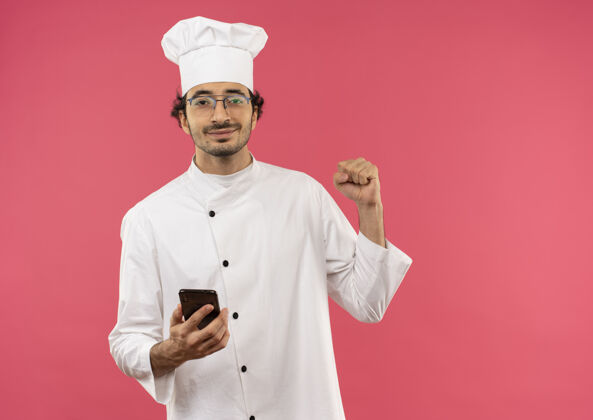 手势年轻的男厨师穿着厨师制服 戴着眼镜 拿着电话 在粉红色的墙上显示“是”的手势电话烹饪手持