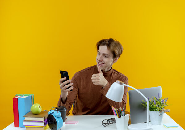 学校微笑的年轻学生男孩坐在书桌旁 手里拿着学习工具 看着电话 他的大拇指孤立地竖起在黄色的墙上年轻人书桌学生