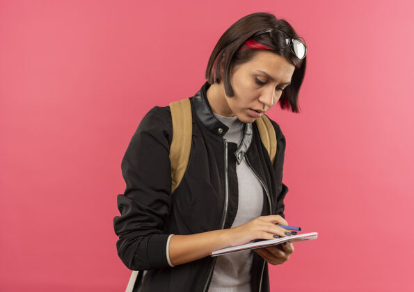 笔记专心的年轻女学生头戴眼镜 背着书包拿着钢笔和笔记本 看着笔记本 把笔记本隔离在粉红色的地方 留着复印空间钢笔空间复制