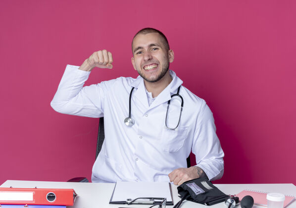 粉色自信的年轻男医生穿着医用长袍和听诊器坐在办公桌旁 手里拿着工作工具 举起拳头孤立在粉红色的桌子上听诊器工作年轻