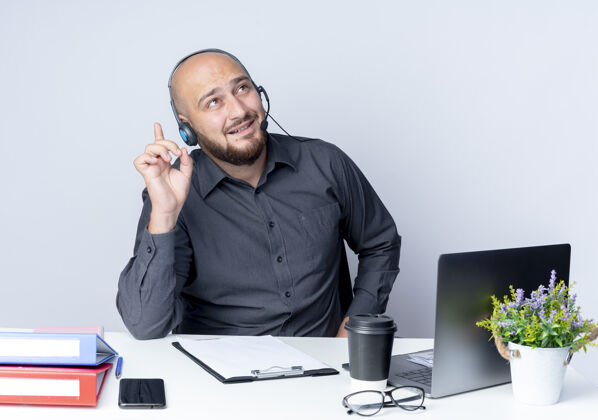 男人令人印象深刻的年轻秃头呼叫中心男子戴着耳机坐在办公桌上 工作工具看着和指着孤立的白色中心印象耳机