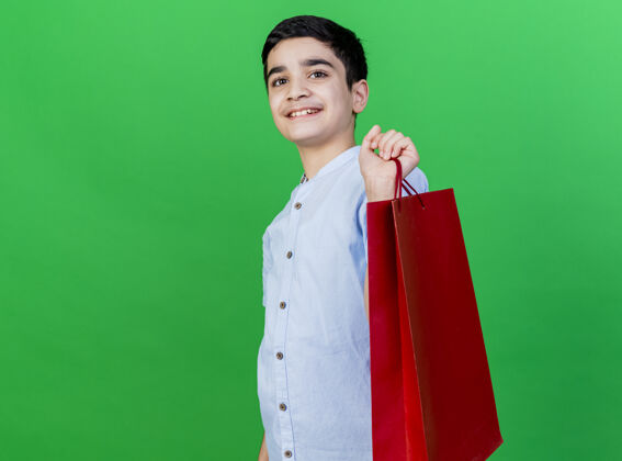 微笑笑容可掬的年轻白人男孩站在侧视图中 手里拿着购物袋 看着隔离在绿色背景上的相机 还有复印空间包相机抱着
