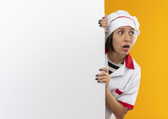 持有令人印象深刻的年轻女厨师身着厨师制服站在后面 手持白色的墙壁 看着橙色的一面孤立的复制空间女性立场壁板