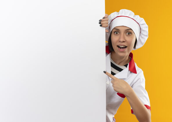 指向穿着厨师制服的年轻女厨师从白色墙壁后面望去 指着橙色的隔离墙 留下了深刻的印象空间印象厨师
