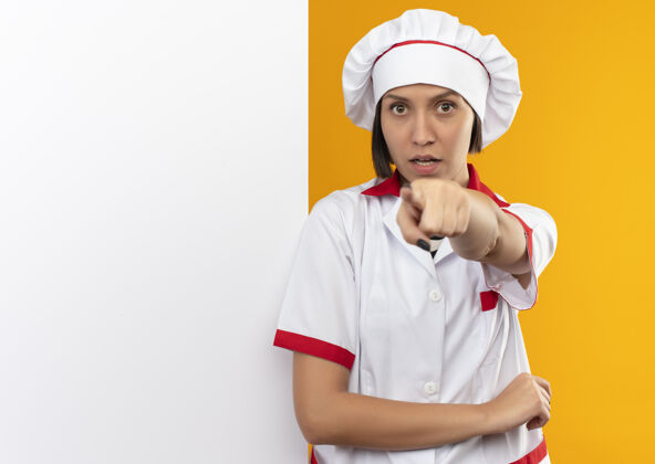 墙壁穿着厨师制服的年轻女厨师站在白色的墙前 孤立地指着橘黄色的复制空间厨师立场烹饪