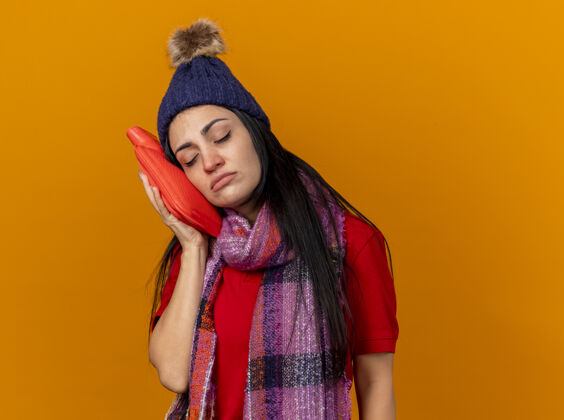 包弱小的年轻白种人患病的女孩戴着冬天的帽子和围巾用热水袋抚摸着脸闭着眼睛隔离在橙色的背景和复制空间围巾封闭女孩