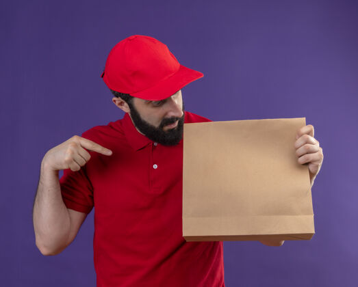 拿印象深刻的年轻英俊的白人送货员身穿红色制服 戴着帽子看着并指着纸包上孤立的紫色紫送红
