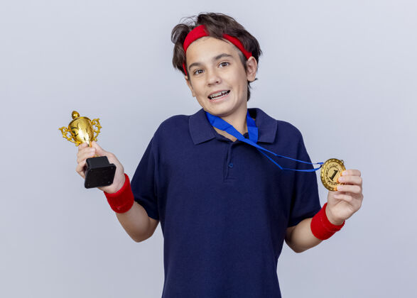 帅气令人印象深刻的年轻英俊的运动男孩戴着头带和护腕 戴着牙套 脖子上戴着奖牌 拿着奖牌和优胜者杯 看着隔离在白色背景上的摄像机牙科背带年轻