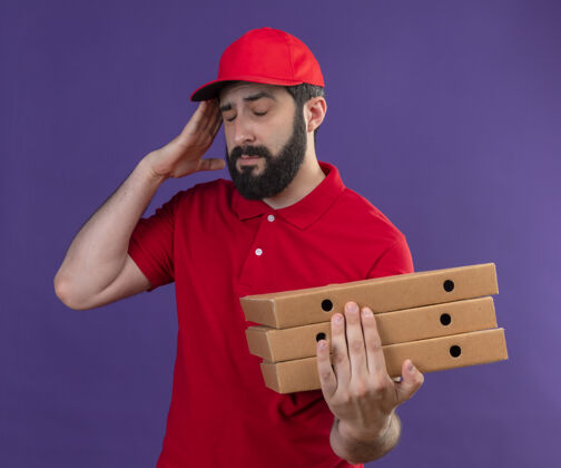 头年轻英俊的白人送货员 身穿红色制服 头戴鸭舌帽 手举披萨盒 闭着眼睛 头痛得要命 孤独地站在紫色的地面上帽子疼痛紫色