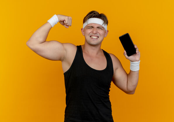 佩戴自信的年轻帅气的运动型男人戴着头带和腕带拿着手机 在橙色上做着强烈的孤立的手势强壮手势运动