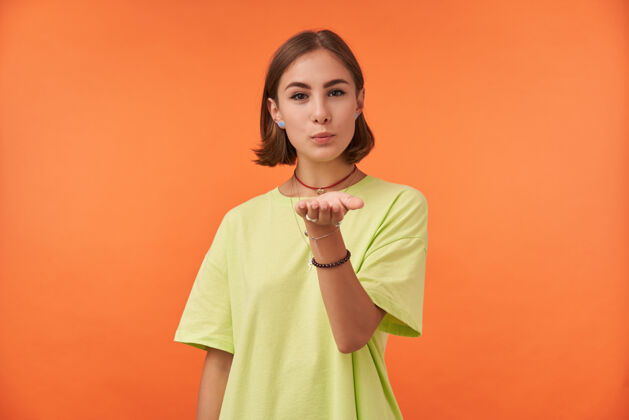 穿孔女学生 一头浅棕色短发的年轻女士 隔着橘色的墙送吻 表示兴趣穿着绿色t恤 项链 手镯和戒指开放成人女人