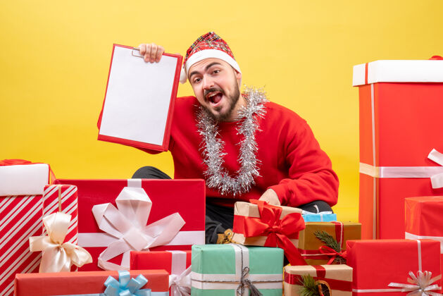 成人正面图：坐在黄色背景上的年轻男性礼物雪圣诞节盒子