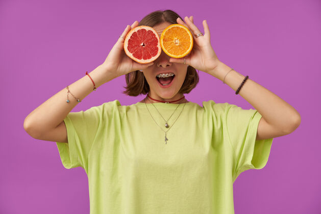 热带女学生 笑容满面的年轻女士 手拿柚子和橘子遮住眼睛 站在紫色的墙上 穿着绿色t恤 戴着牙套 手镯和项链女性时髦深色
