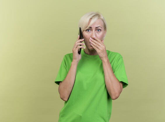 电话惊讶的中年金发斯拉夫女人在电话里说话 看着相机 手放在嘴上 在橄榄绿的背景上 有复印空间惊喜金发保持