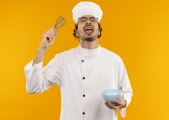 男性年轻快乐的男厨师闭着眼睛 穿着厨师制服 戴着玻璃杯 拿着碗 在黄色的墙上露出孤立的舌头眼镜喜悦制服