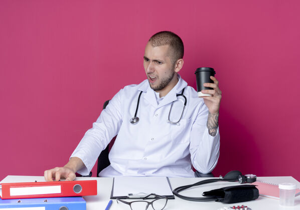 桌子恼怒的年轻男医生穿着医用长袍和听诊器坐在办公桌旁 手里拿着工作工具 手里拿着塑料咖啡杯 孤立地看着粉红色的办公桌长袍咖啡听诊器
