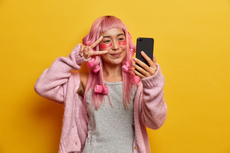 粉色满头粉红头发的快乐亚洲女人 在眼睛上做和平手势 自拍 在眼睛下面贴胶原蛋白贴片垫子美丽眼睛