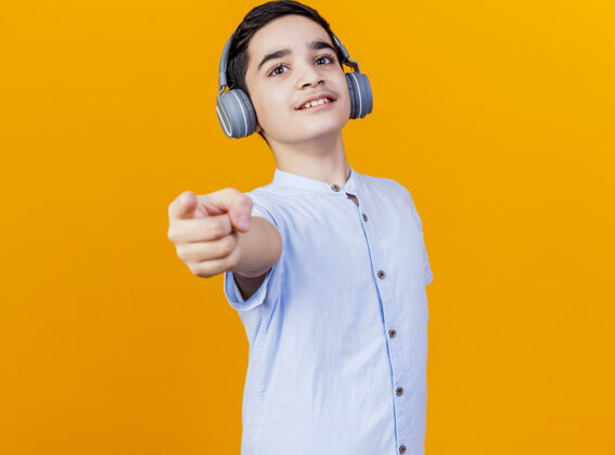 复制高兴的年轻白人男孩戴着耳机看着和指着相机隔离在橙色背景与复制空间空间年轻耳机