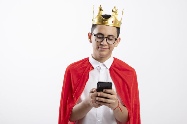 使用高兴的年轻超级英雄男孩在红斗篷戴眼镜和皇冠用手机隔离在白色背景与复制空间皇冠请手机