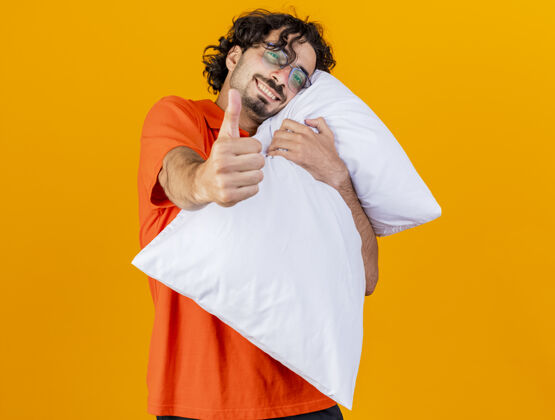 年轻带着微笑的年轻白种人 戴着眼镜 抱着枕头 看着相机 在橙色背景上孤立地竖起大拇指 留着复印空间拇指显示枕头