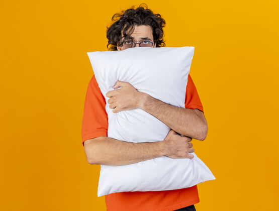男人弱小的年轻白种人 戴着眼镜 抱着枕头 从后面看相机 橙色背景 有复印空间枕头橙色疾病