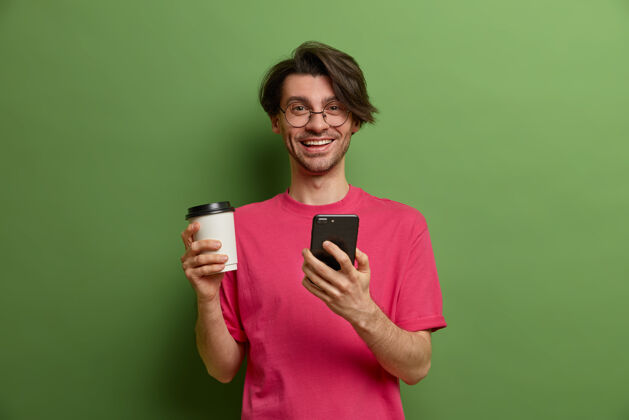 杯子快乐微笑的家伙在网上商店里搜索必需品 使用智能手机应用程序 浏览社交网络 从纸杯里喝芳香的咖啡 有时髦的发型 在室内摆姿势在线男性年轻