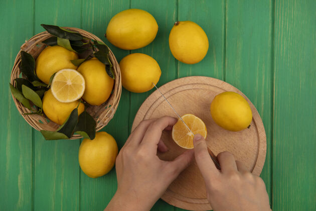 食物俯视图中的女性手切割柠檬在一个木制的厨房板上用刀与柠檬桶在一个绿色的木墙上板新鲜柑橘
