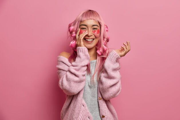 人类开朗的亚洲女孩肖像使用具有抗皱效果的水凝胶贴片 粉色头发上戴卷发器 真诚微笑 穿着休闲套头衫成人牙齿粉色头发