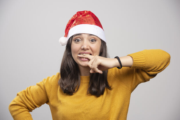 女性戴圣诞帽的女人在灰色的墙上咬手指帽子礼品盒女士