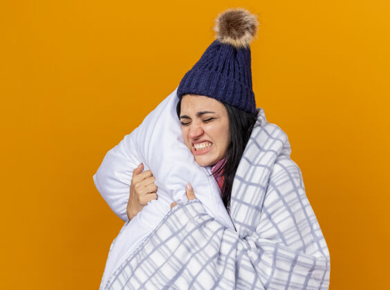 眼睛疼痛的年轻白种人生病的女孩 戴着冬天的帽子和围巾 裹在格子布抱枕里 闭着眼睛 隔离在橙色背景上 留着复印空间穿着格子围巾