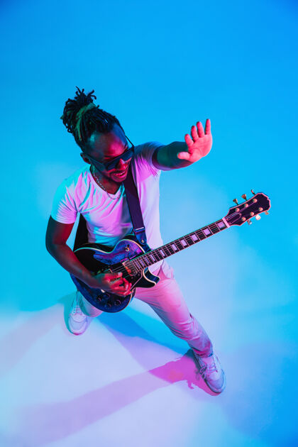 帅气年轻的非洲裔美国音乐家在霓虹灯下像摇滚明星一样在蓝色背景下弹吉他旋律立场复古
