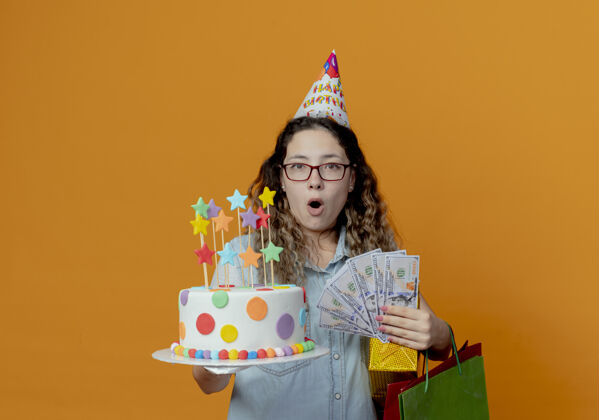 惊喜惊讶的年轻女孩戴着眼镜 戴着生日帽 手里拿着生日蛋糕 手里拿着礼盒 袋子和钱放在橘子上礼物蛋糕钱