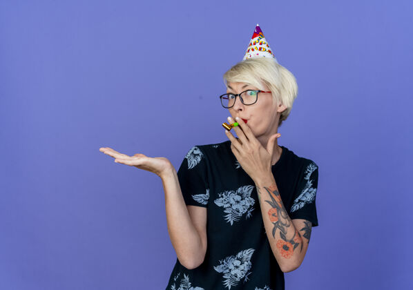 人给人印象深刻的年轻金发派对女郎戴着眼镜和生日帽看着前面拿着派对吹风机嘴里露出空手孤立在紫色的墙上衣服聚会生日