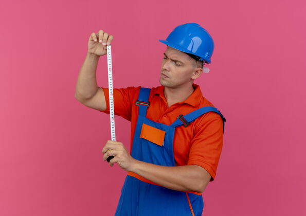 思考想着年轻的男建筑工人穿着制服 戴着安全帽 拿着并看着粉色的仪表胶带米制服胶带